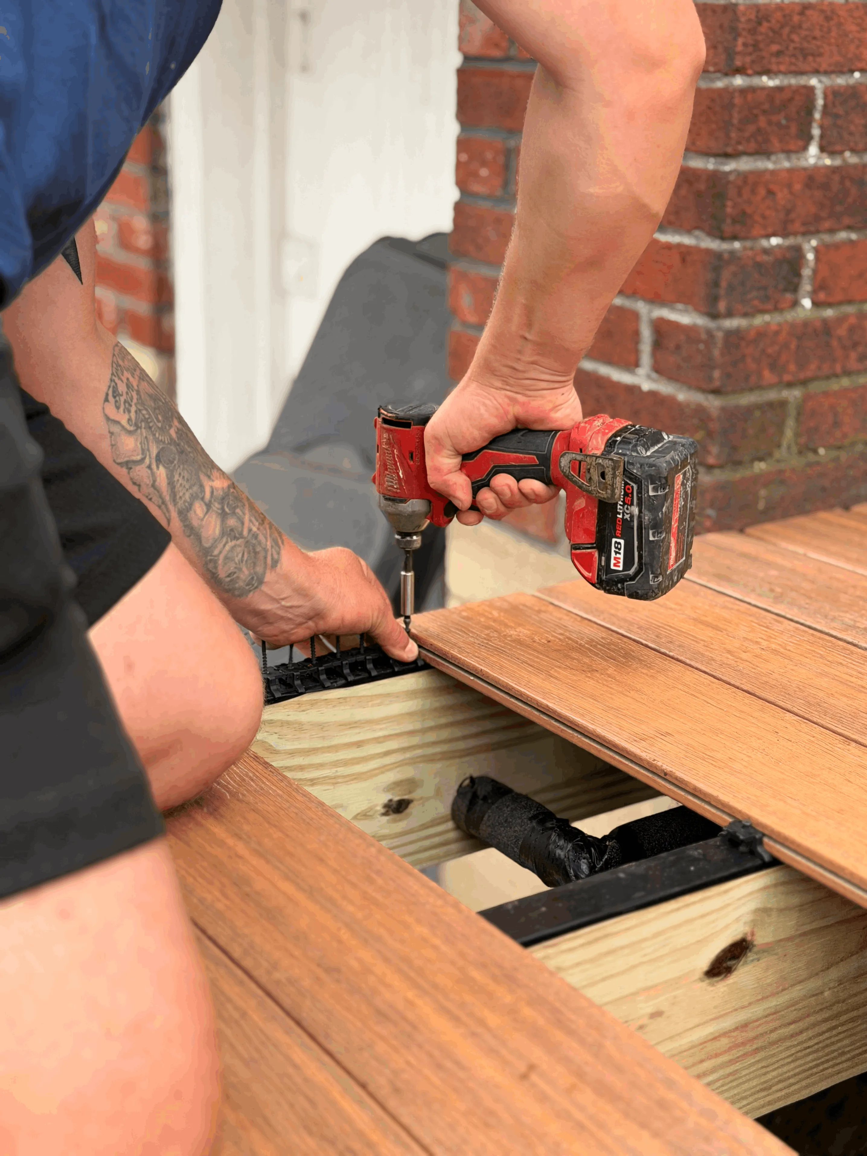 Technician fastening a composite deck board alongside a brick wall