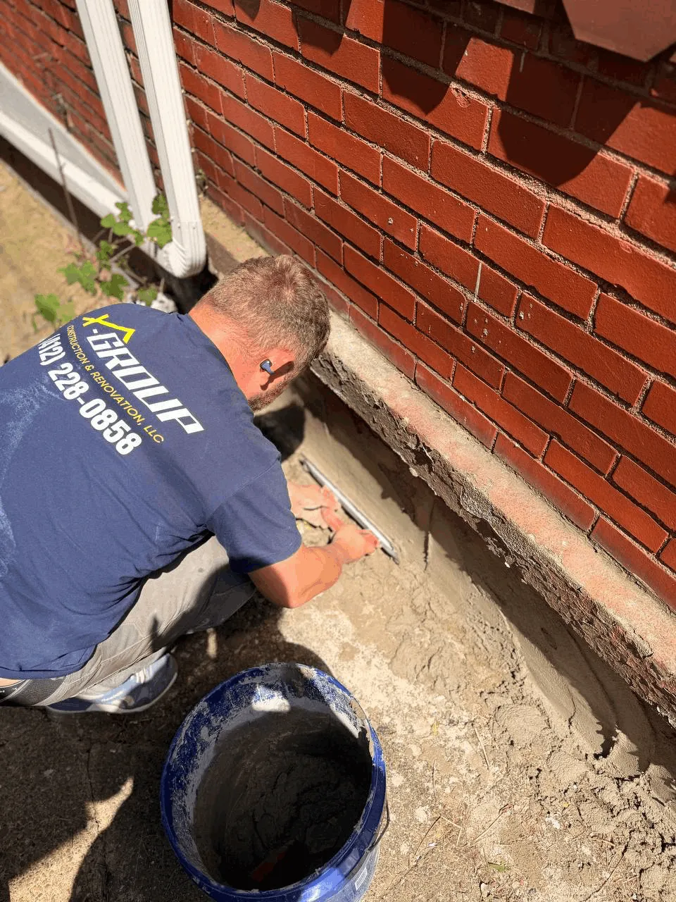 X-Group technician finishing concrete along a brick foundation wall