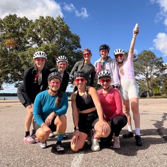The gang posing under a blue sky, one rider cheering with her water bottle in the air