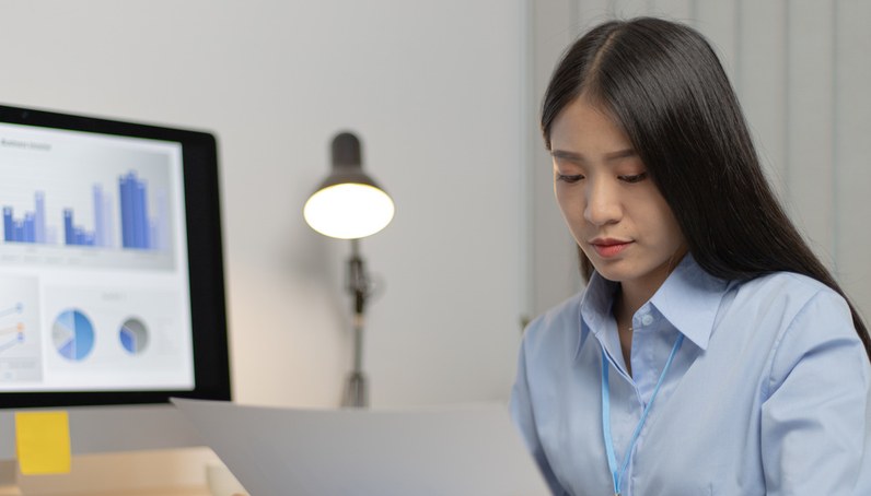 Young woman in a blue shirt reviewing a document at her desk with a computer screen displaying charts and graphs in the background.