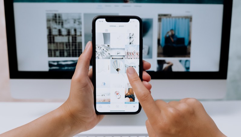 Person holding a smartphone with a photo gallery app open, browsing images in a grid, in front of a blurred desktop computer screen.