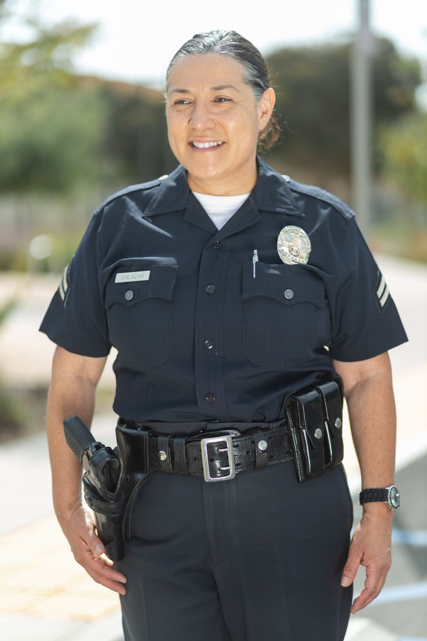 A smiling police officer in uniform standing outdoors on a sunny day.