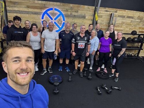 Group of people smiling in a gym with weights on the floor and wood panel wall with blue logo behind.