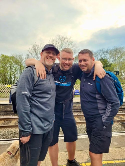 Three men smiling and posing together on a train station platform with a small dog nearby, all friends from the gym.