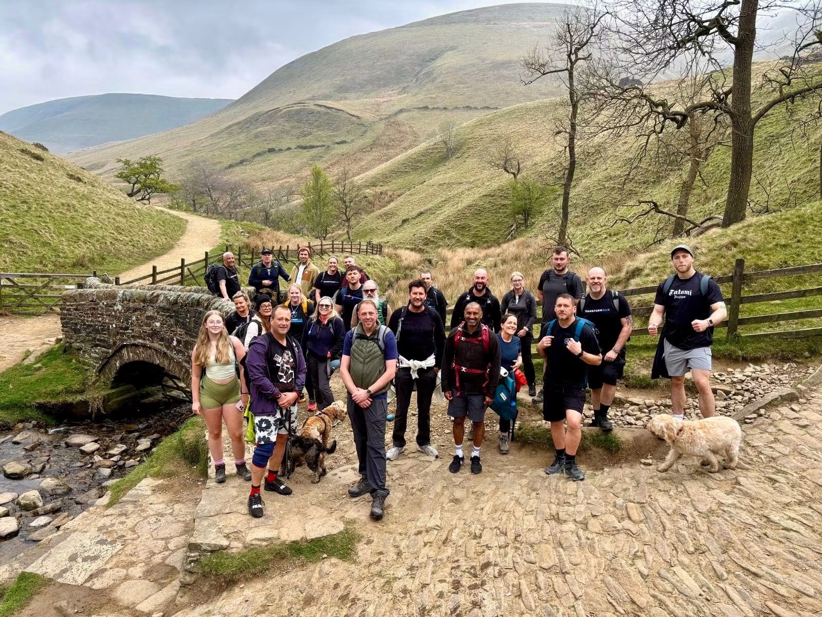 Gym community of hikers with backpacks and two dogs posing near a stone bridge and grassy hills.