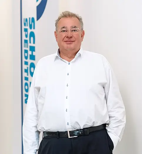 Matthias Schott steht mit weißem Hemd und dunkler Hose vor einer Wand mit Fahnen mit dem Schriftzug 'SCHOTT SPEDITION'.
