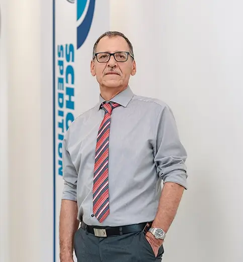 Harald Rhein steht mit weißem Hemd und dunkler Hose vor einer Wand mit Fahnen mit dem Schriftzug 'SCHOTT SPEDITION'.