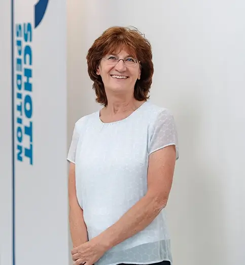 Birgit Schatz-Walter steht mit weißer Bluse und dunkler Hose vor einer Wand mit Fahnen mit dem Schriftzug 'SCHOTT SPEDITION'.