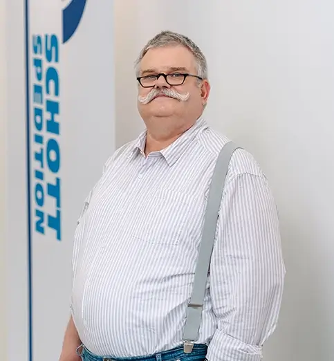 Thomas Funk steht mit weißem Hemd und dunkler Hose vor einer Wand mit Fahnen mit dem Schriftzug 'SCHOTT SPEDITION'.