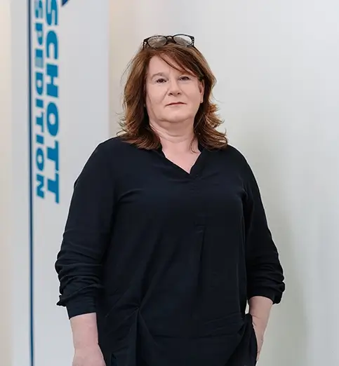 Regina Bartels steht mit dunkler Bluse und dunkler Hose vor einer Wand mit Fahnen mit dem Schriftzug 'SCHOTT SPEDITION'.
