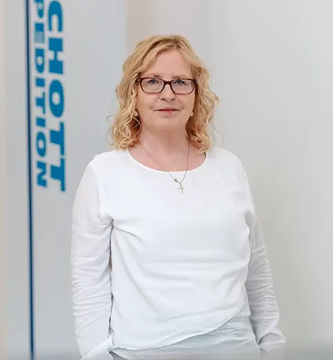 Martina Hiesch steht mit heller Bluse und dunkler Hose vor einer Wand mit Fahnen mit dem Schriftzug 'SCHOTT SPEDITION'.