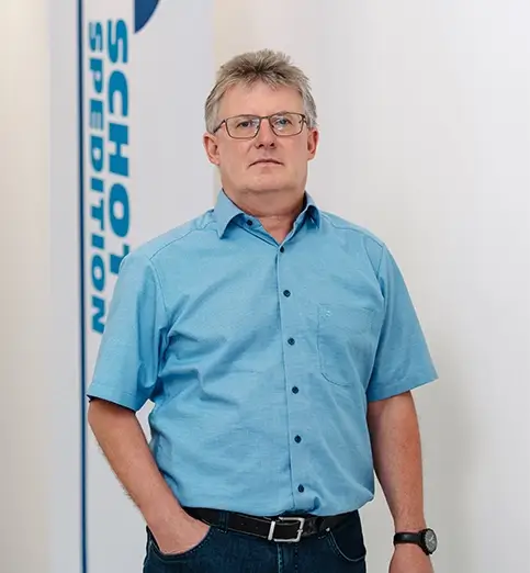 Stefan Russ steht mit blauem Hemd und dunkler Hose vor einer Wand mit Fahnen mit dem Schriftzug 'SCHOTT SPEDITION'.