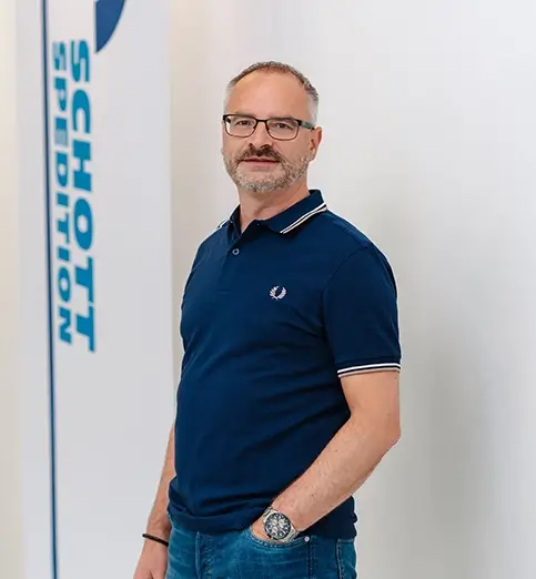 Mario Fischer steht mit dunklem Shirt und dunkler Hose vor einer Wand mit Fahnen mit dem Schriftzug 'SCHOTT SPEDITION'.
