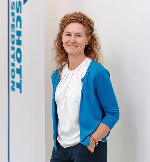 Gudrun Lauchs steht mit heller Bluse und dunkler Hose vor einer Wand mit Fahnen mit dem Schriftzug 'SCHOTT SPEDITION'.