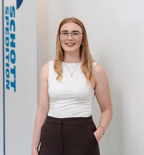 Julia Hofmann steht mit heller Bluse und dunkler Hose vor einer Wand mit Fahnen mit dem Schriftzug 'SCHOTT SPEDITION'.