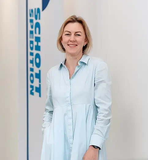 Oleksandra Petukh steht mit heller Bluse und dunkler Hose vor einer Wand mit Fahnen mit dem Schriftzug 'SCHOTT SPEDITION'.