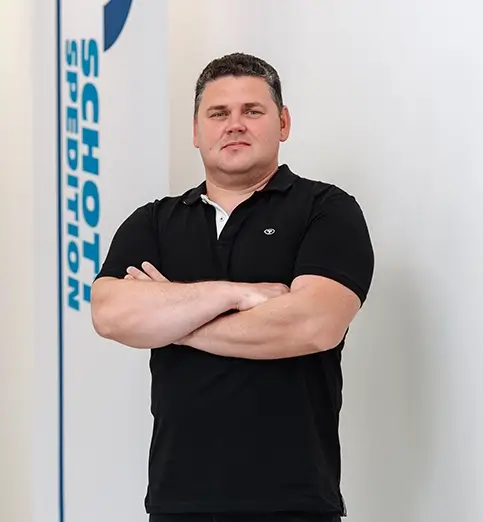 Alexander Mokan steht mit dunklem Shirt und dunkler Hose vor einer Wand mit Fahnen mit dem Schriftzug 'SCHOTT SPEDITION'.