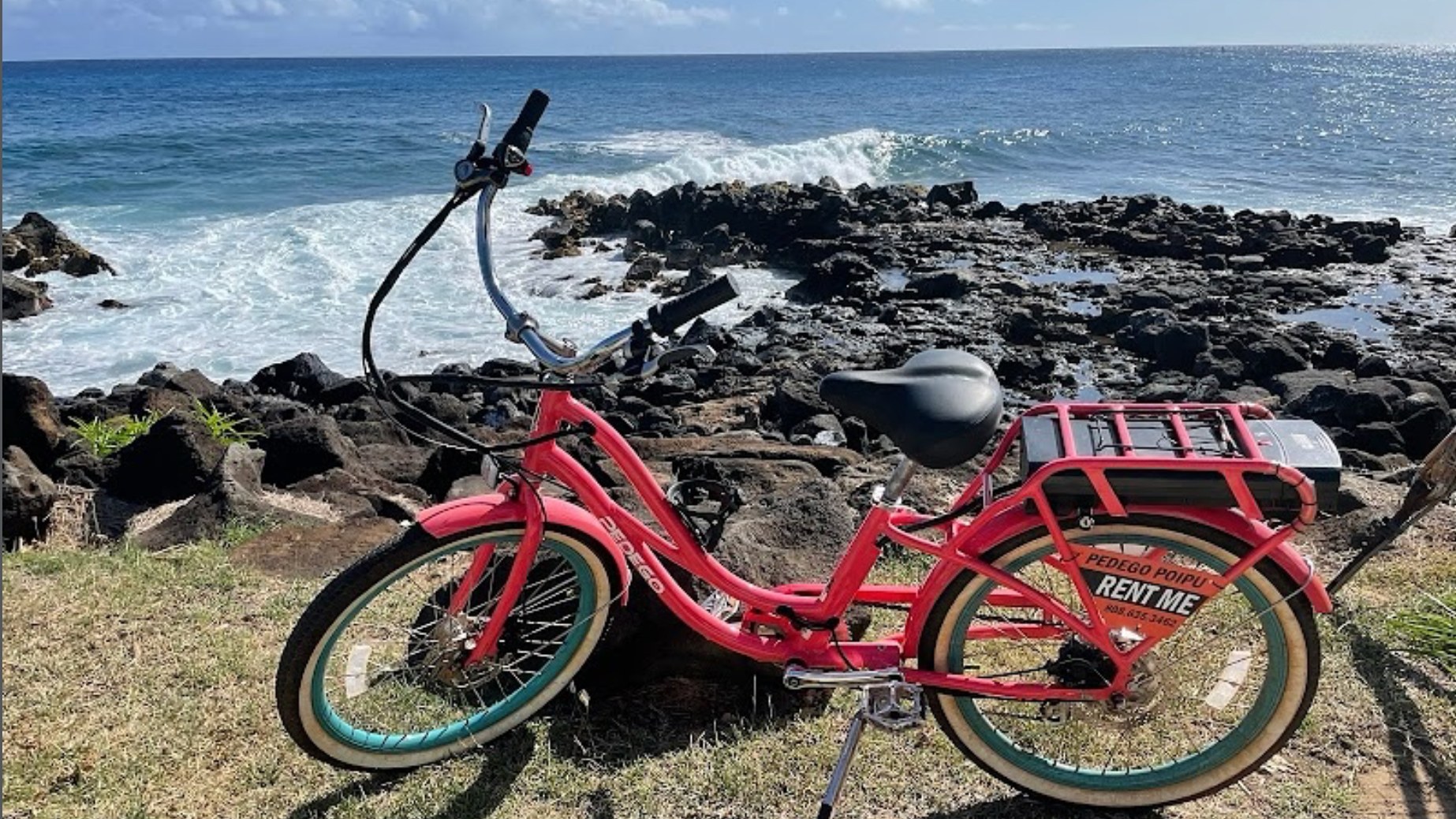 A coral Pedego electric cruiser parked on the grass above a rocky stretch of the Poipu coastline, with surf breaking behind it