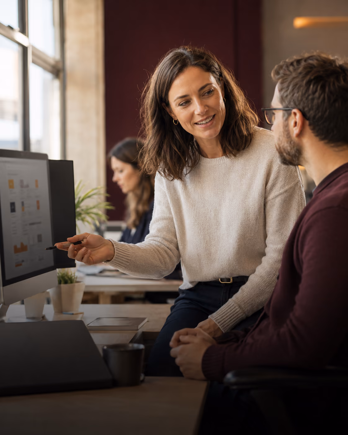 A woman in a white sweater gestures with a pen while talking to a seated man wearing glasses in a modern office setting, with a computer monitor showing charts and a woman working in the background.