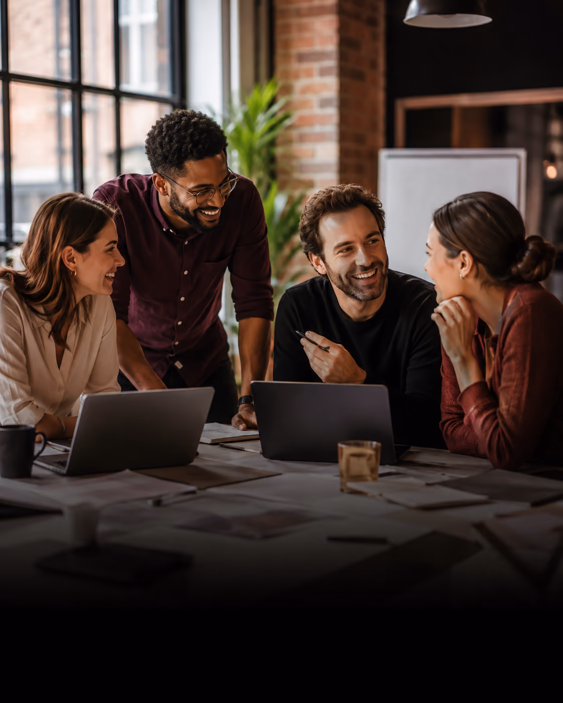 Four diverse colleagues engaged in a lively discussion around a table with laptops and papers in a modern office with large windows and exposed brick walls.