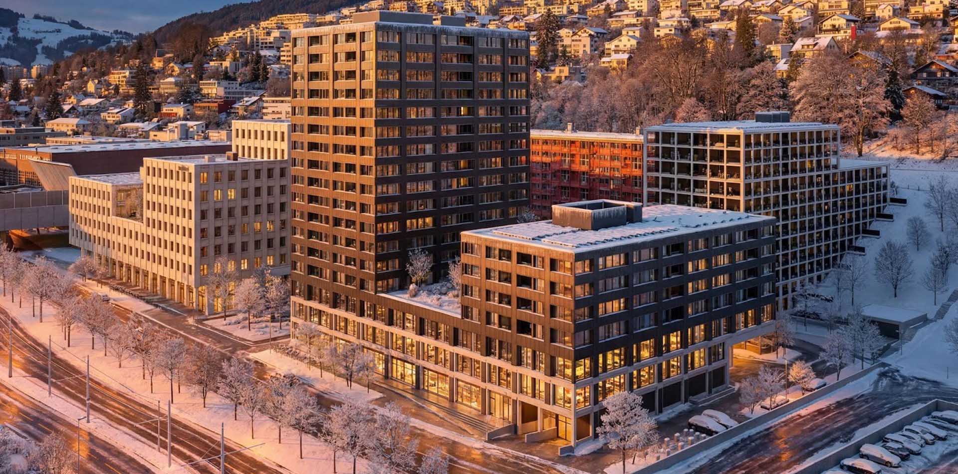 Modern multi-story office buildings with large windows lit warmly from inside, situated in a snowy urban landscape at sunset, with snow-covered roads, parked cars, trees, and a hillside with residential houses in the background.