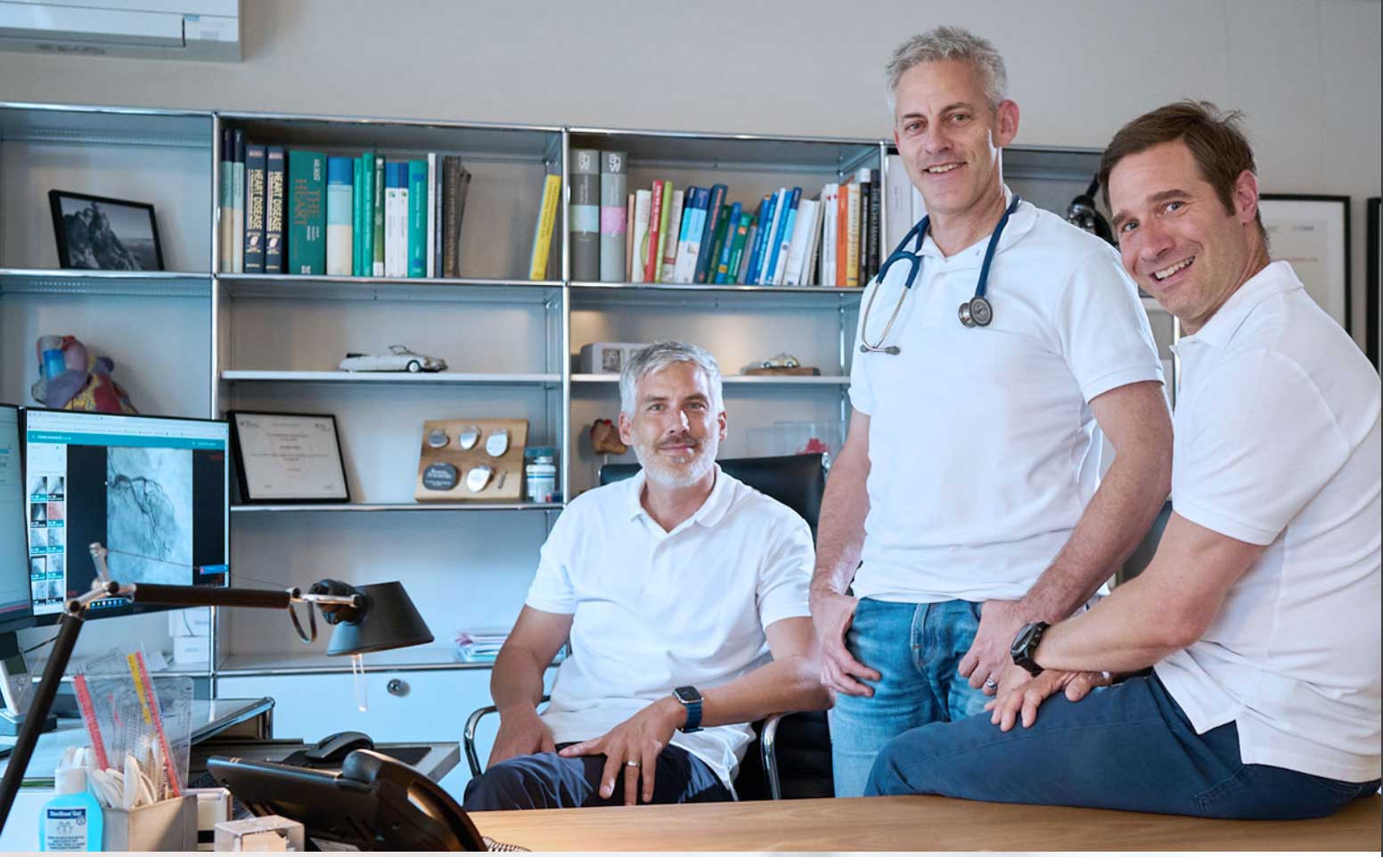 Three male doctors in white polo shirts in a medical office, with one seated at a desk and two standing nearby, one wearing a stethoscope around his neck.