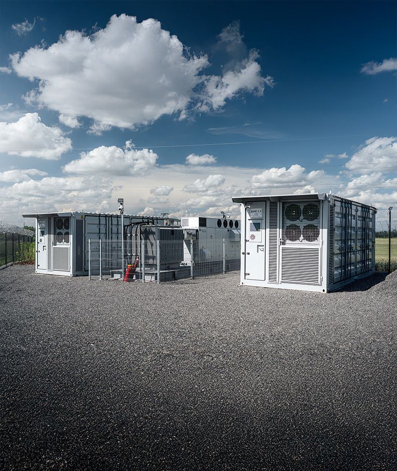Dva technologické kontejnery bateriového úložiště TESLA Energy Group na štěrkové ploše pod modrou oblohou