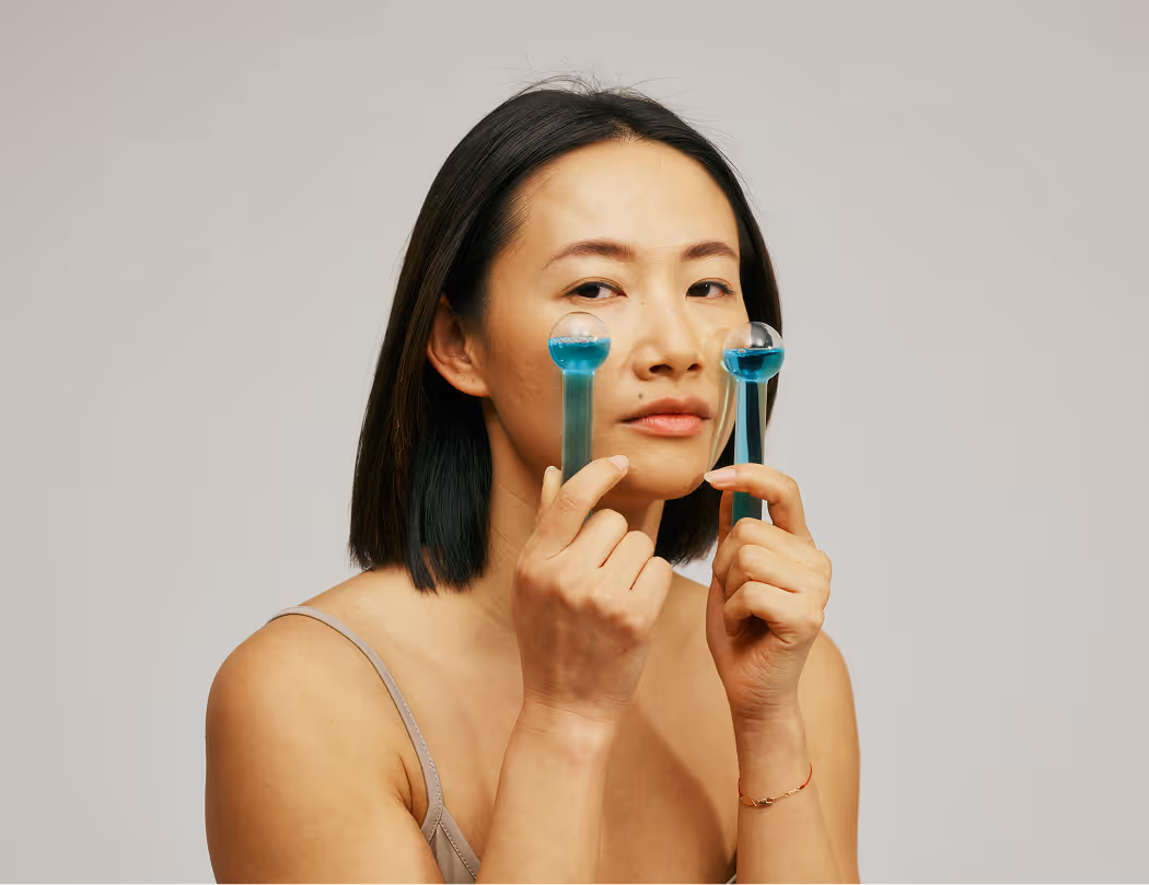 Woman using two blue facial massage rollers near her eyes against a neutral background.