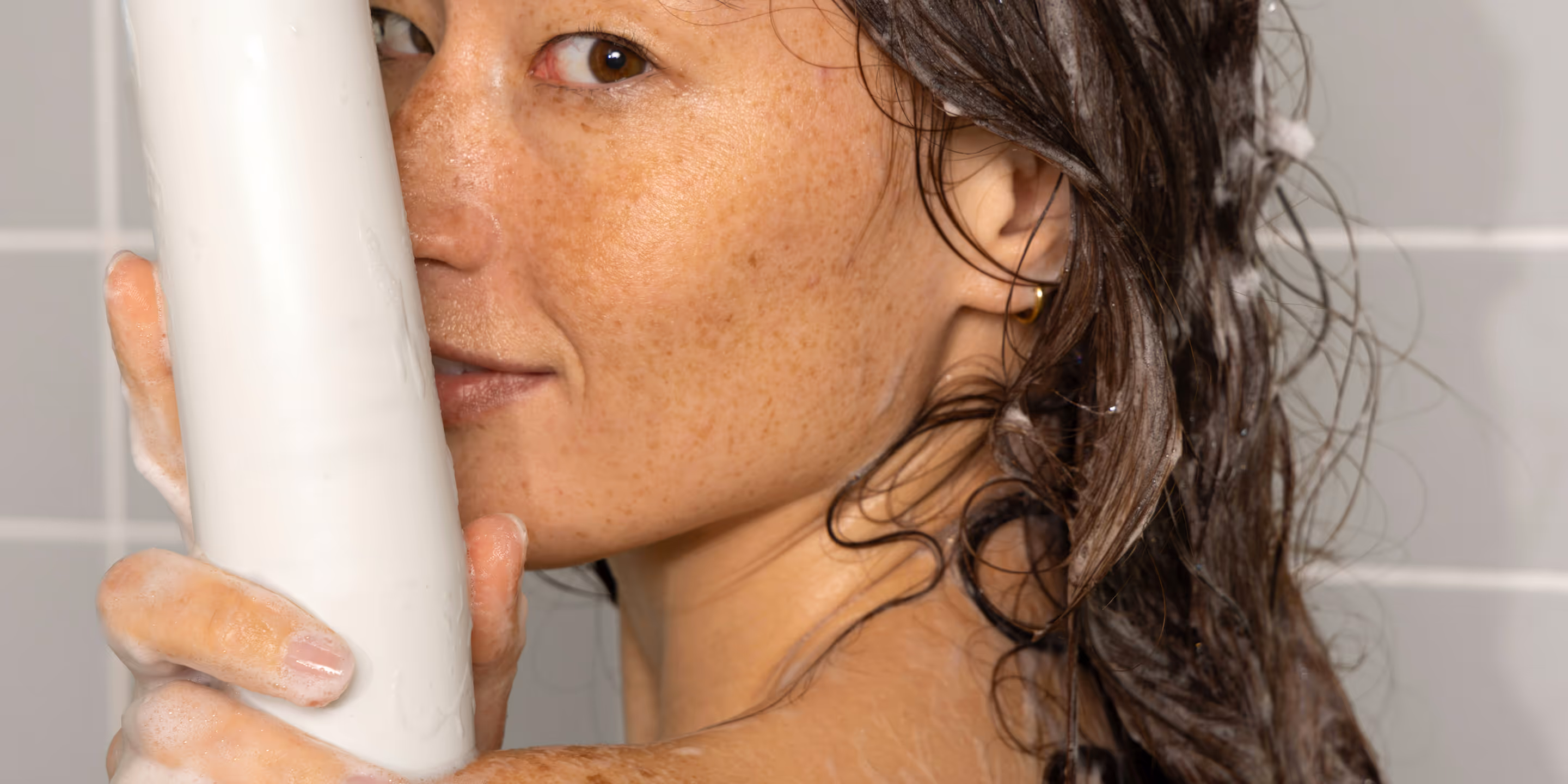 Woman with wet hair holding a large white bar of soap close to her face in a tiled shower.