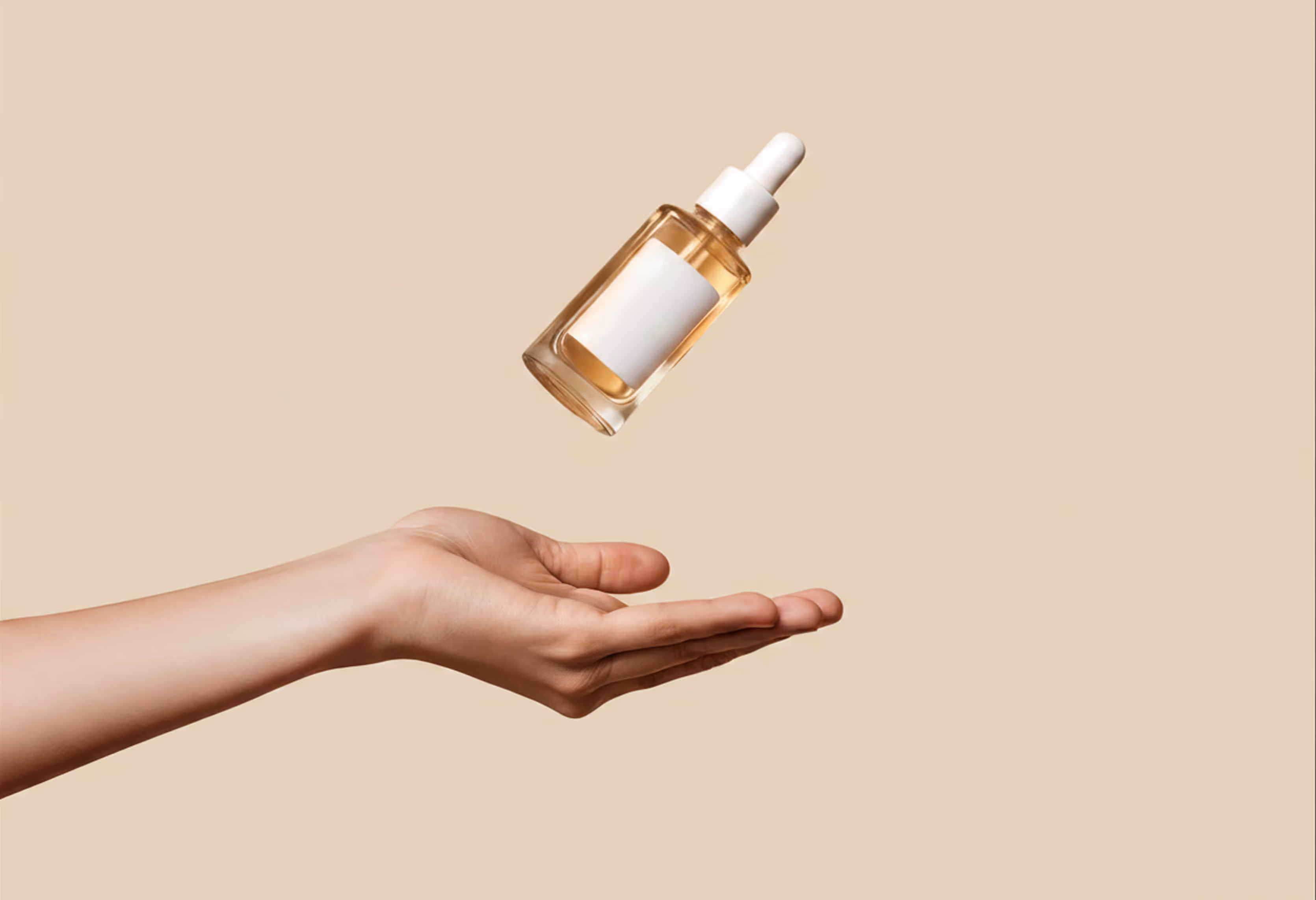Hand with palm open below a floating glass dropper bottle filled with light golden serum against beige background.