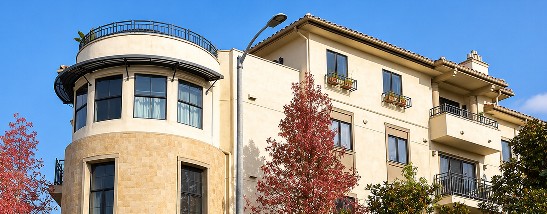 Modern three-story apartment building with beige and tan exterior, multiple windows, balconies with metal railings, and red-leaved trees in front under a clear blue sky.