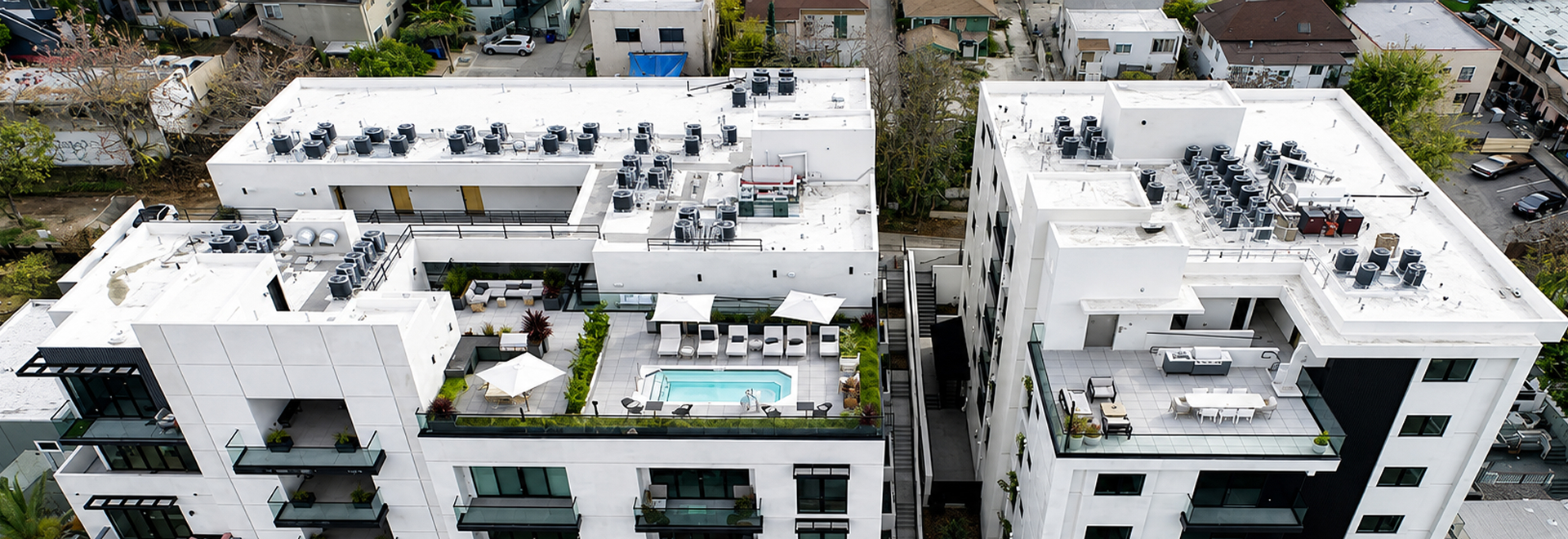 Aerial view of two modern white apartment buildings with rooftop amenities including a small swimming pool surrounded by lounge chairs, umbrellas, outdoor seating areas, and greenery, situated in a residential neighborhood.