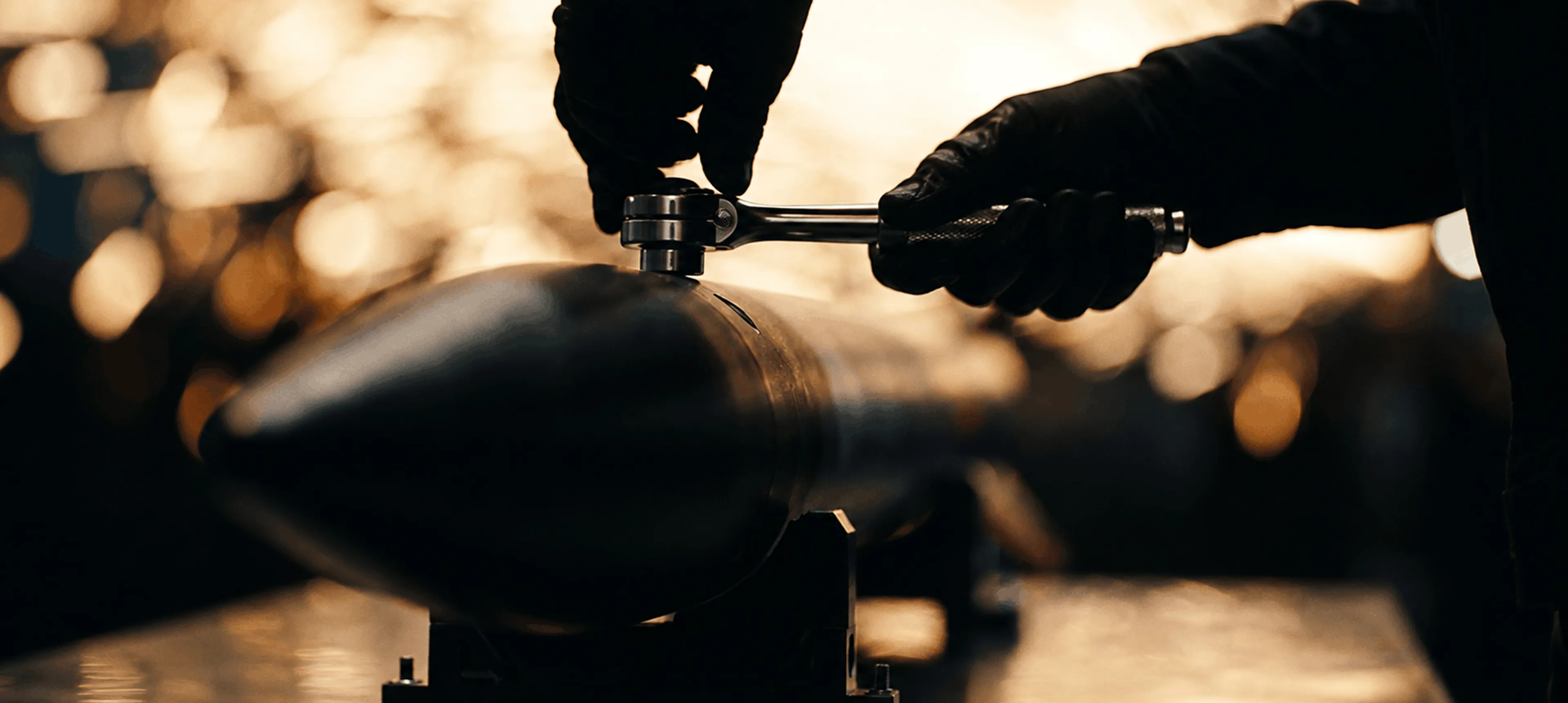 Hands wearing black gloves using a wrench to tighten or loosen a bolt on a large cylindrical metallic object with a pointed end, against a blurred warm-toned background.
