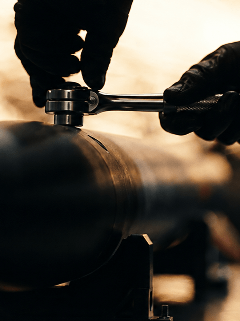 Close-up of a person wearing black gloves using a ratchet wrench to tighten or loosen a bolt on a large cylindrical metal pipe.