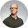 Portrait of a bald man with glasses and a green sweater smiling against a gray background.