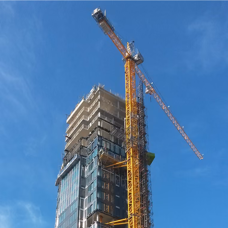 Tower crane lifting facade units alongside a high-rise under construction against a blue sky.