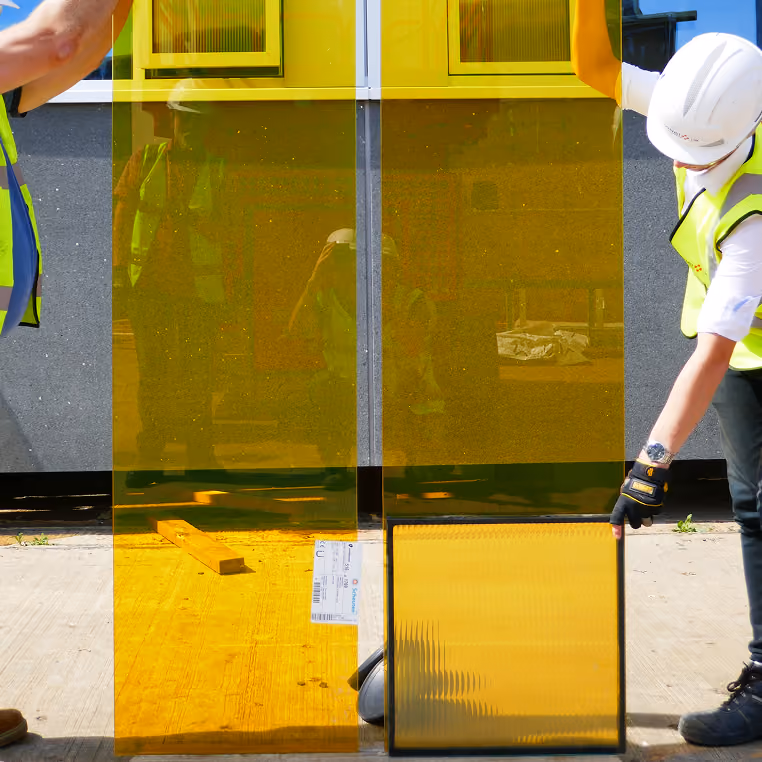 Workers in hi-vis vests and helmets handling amber-tinted insulating glass units during mock-up testing.