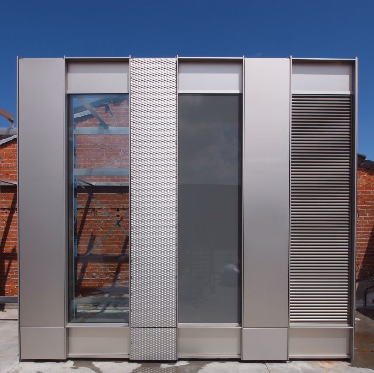 Facade mock-up panel combining perforated metal, louvres and glazing, mounted in front of a brick building.