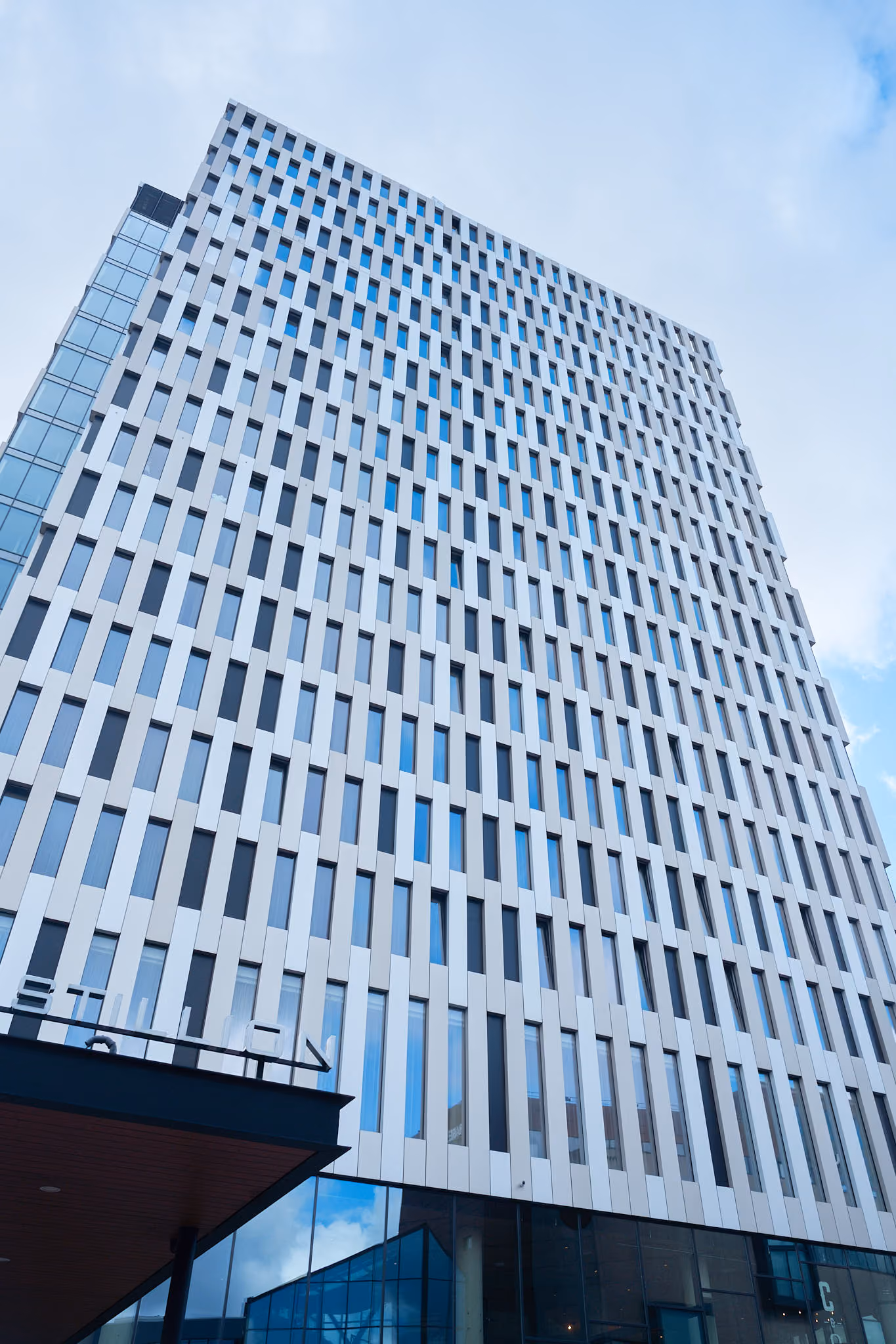 Modern high-rise office building with a pattern of vertical white and glass panels under a partly cloudy blue sky.