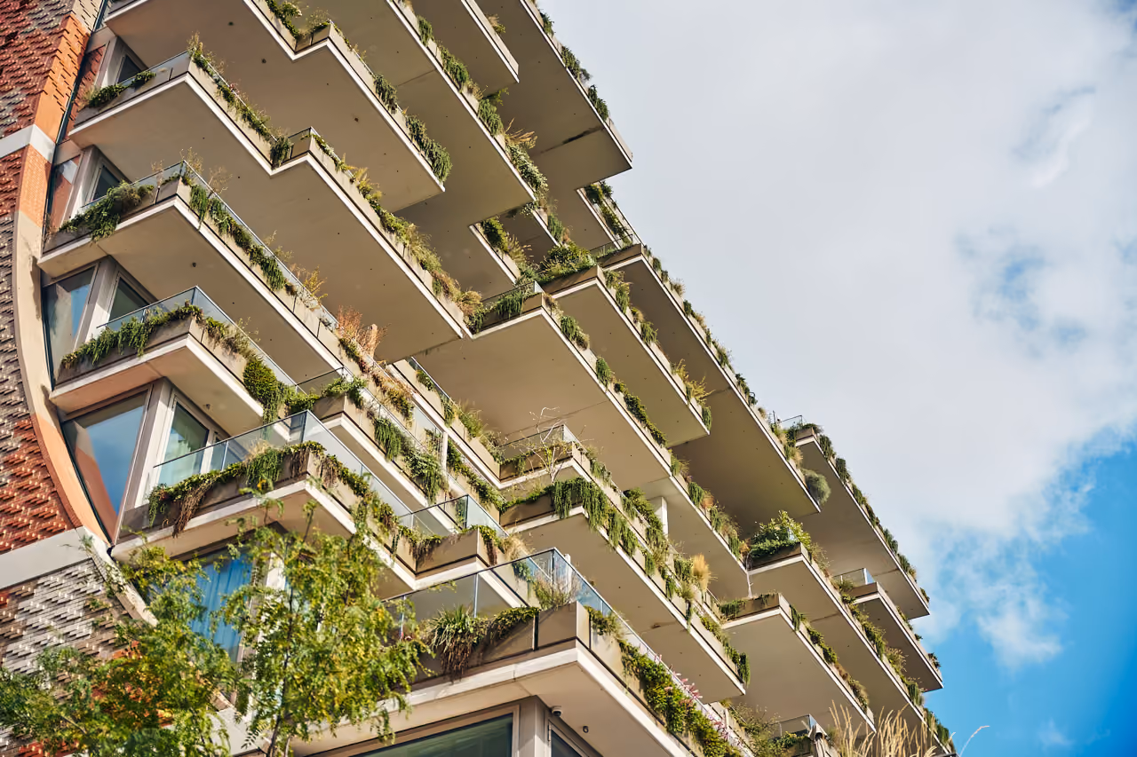 Modern multi-storey building viewed from below with staggered balconies adorned with abundant green plants and shrubs against a partly cloudy blue sky.