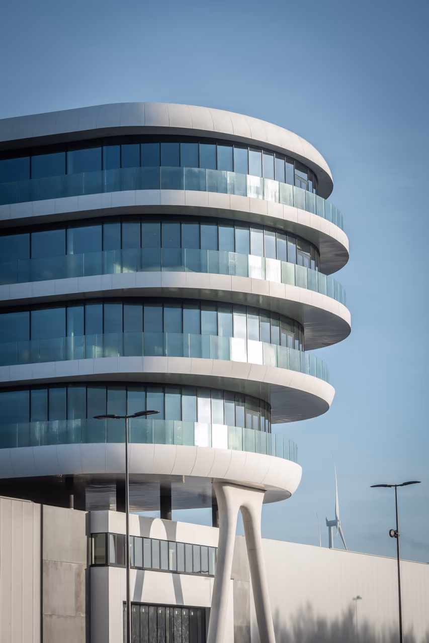 Modern building with rounded balconies and glass railings stacked in circular layers against a clear blue sky, featuring sleek supporting pillars below and wind turbines visible in the background.