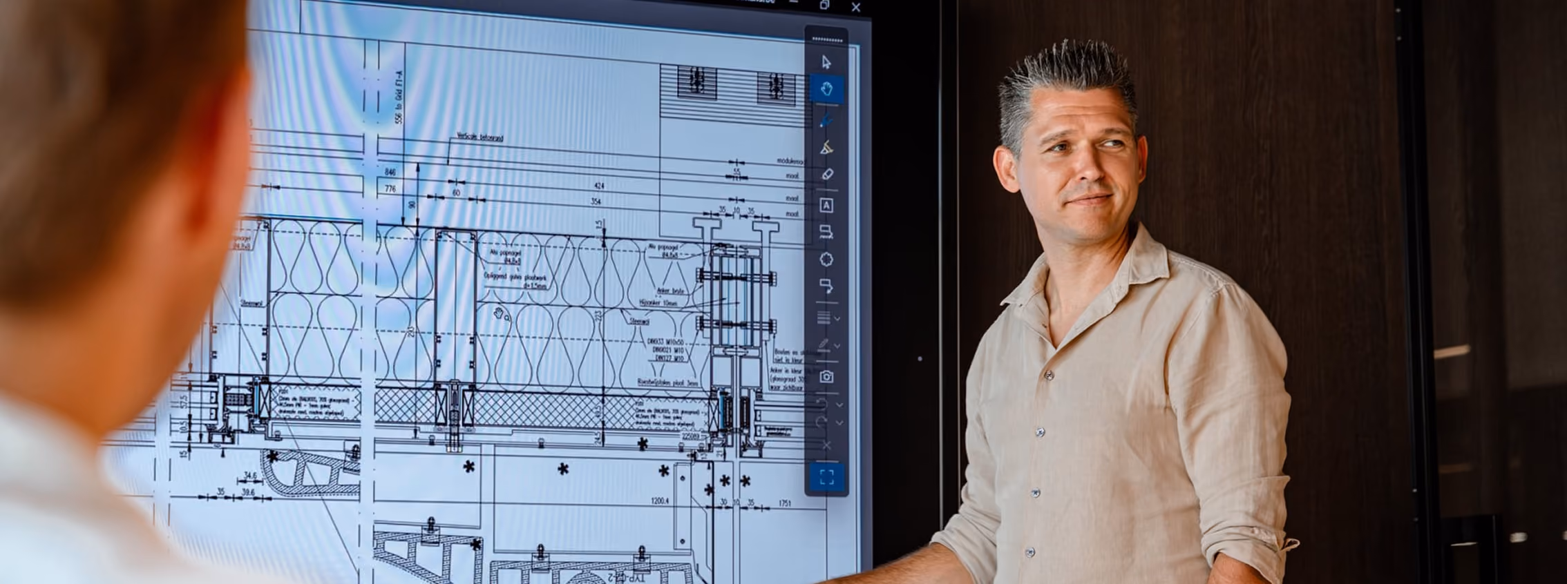 Man in a beige shirt presenting architectural blueprints on a large digital screen to another person in an office setting.