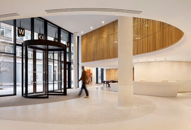 Spacious modern building lobby with large glass revolving door, wooden slatted upper wall feature, white reception desk, and person walking inside.