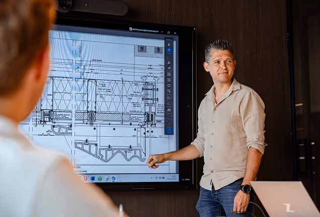 Man in casual shirt and jeans giving a presentation in a meeting room, pointing at architectural blueprints displayed on a large screen while two colleagues listen.