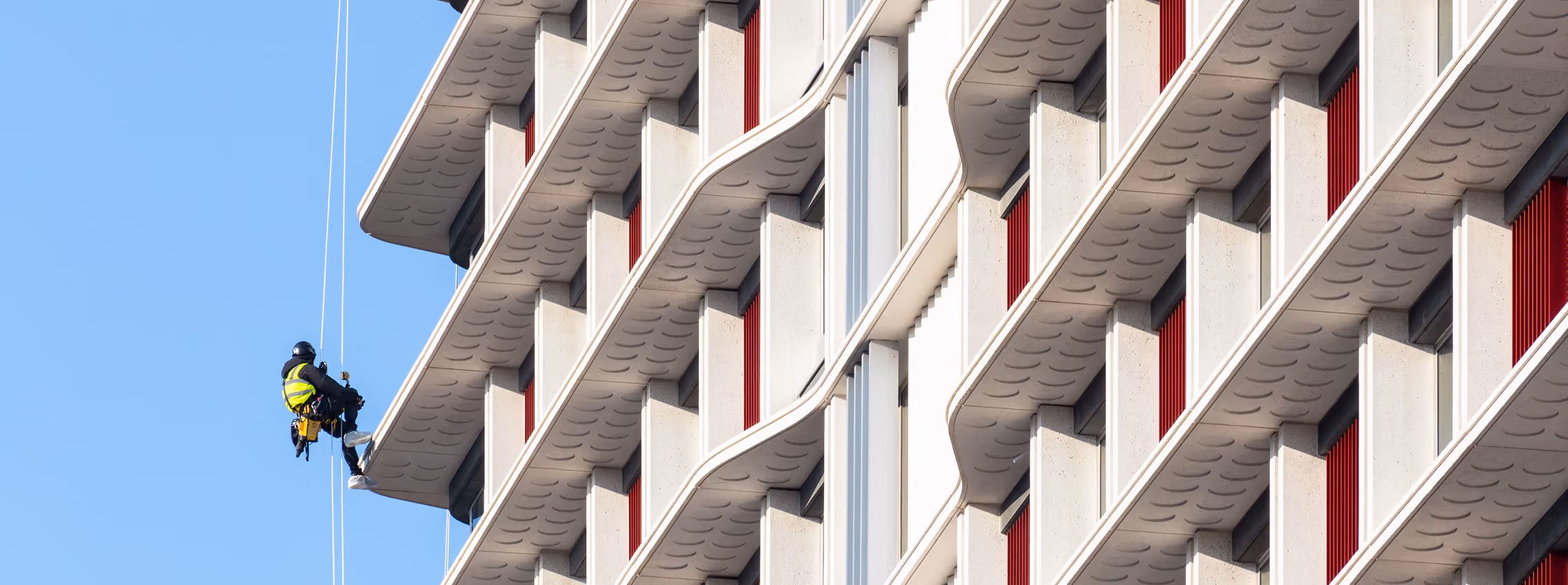 Worker wearing safety harness and bright jacket suspended by ropes cleaning or inspecting the exterior facade of a modern building with repetitive geometric balconies and red window shutters.