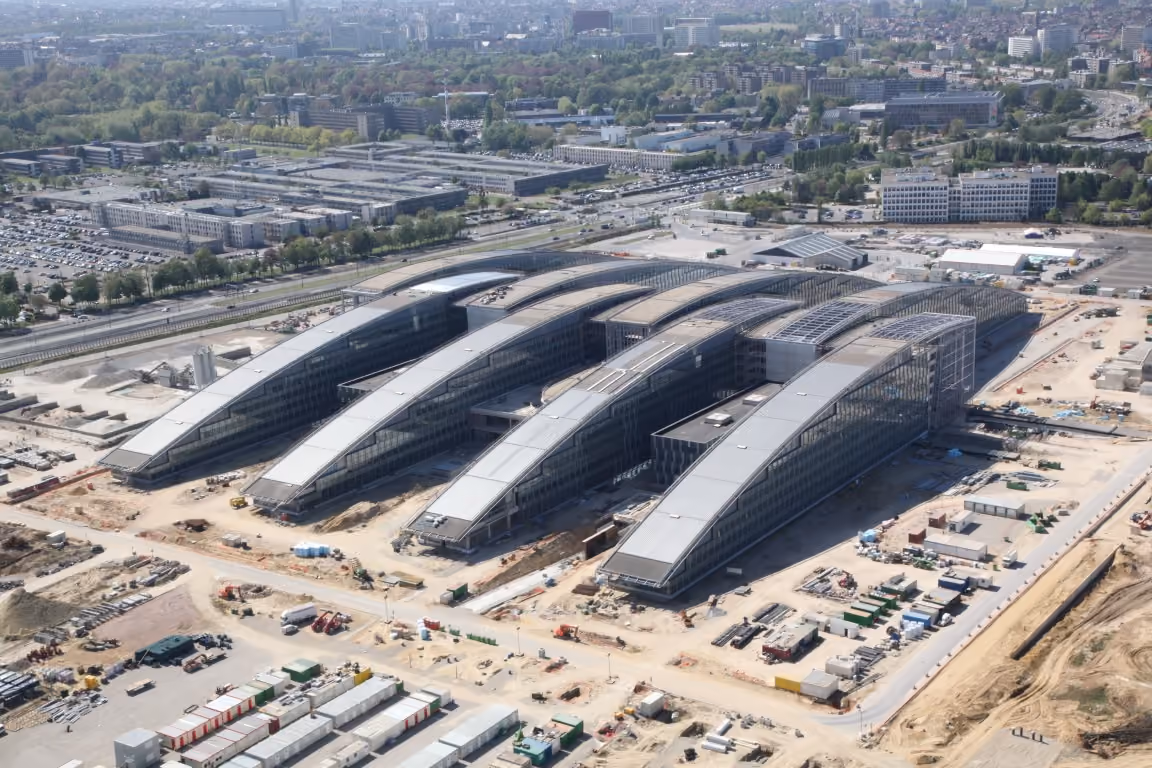 Aerial view of four large curved glass buildings under construction with surrounding construction equipment and materials, located near roads and greenery in an urban area.