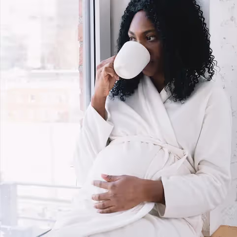 Pregnant woman sitting in a white robe, drinking from a cup and considering probiotics during pregnancy