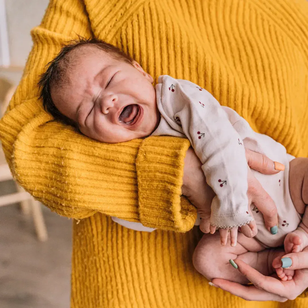 Crying baby being held by an adult in a yellow sweater, to represent concerns about the effects of antibiotics on infants