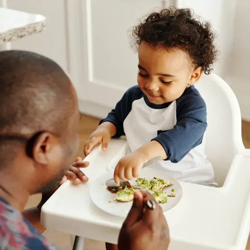 A father feeds his toddler in a high chair, introducing solid foods