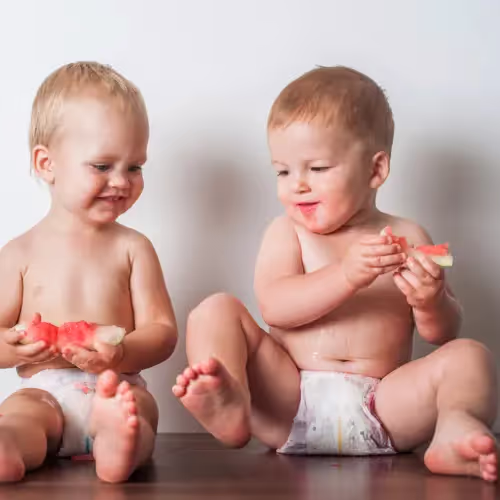 Two babies in diapers sit together happily eating watermelon